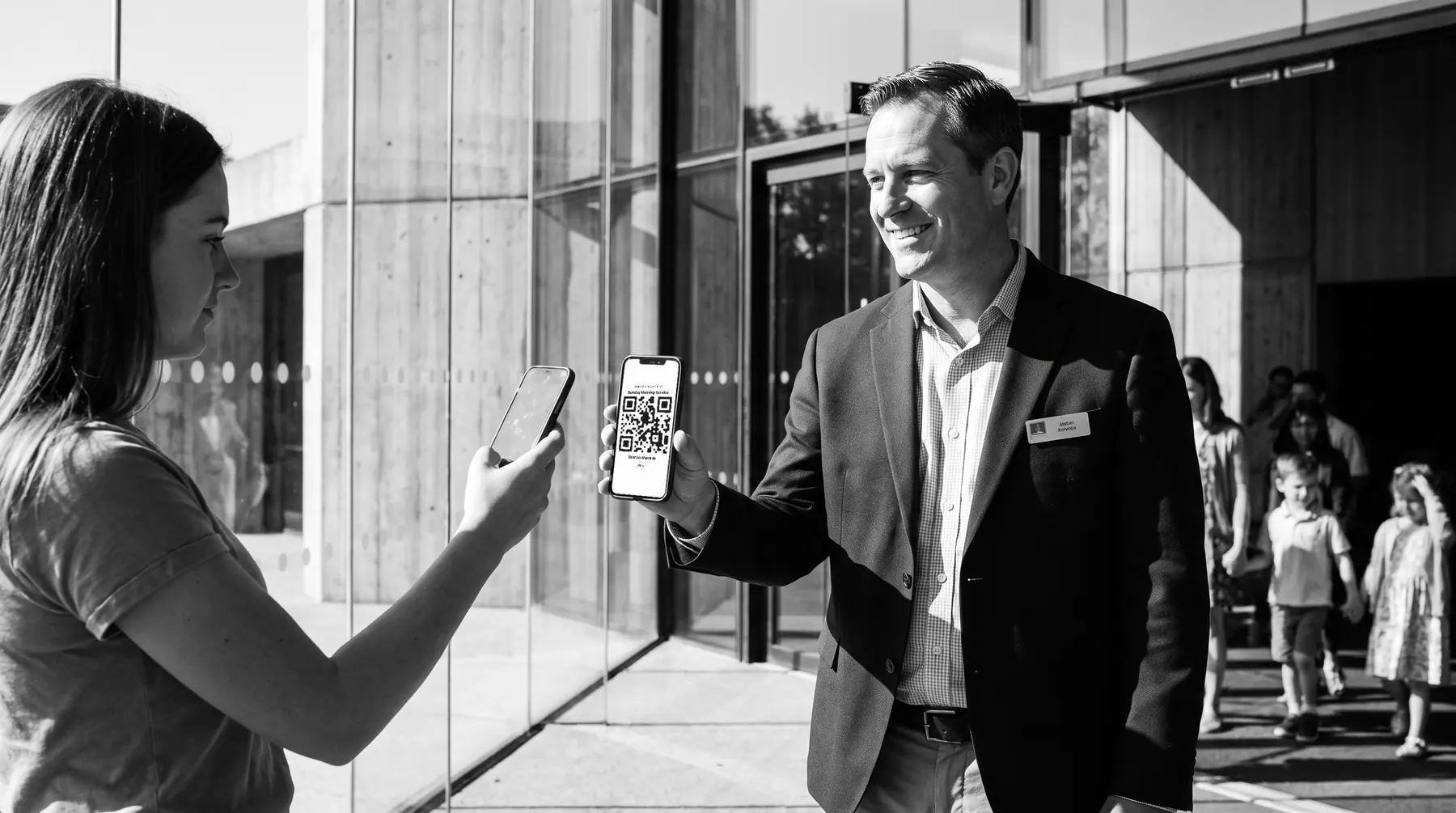 At the church entrance, an usher holds up his phone showing the Attend check-in QR code while an arriving member scans it with her own phone